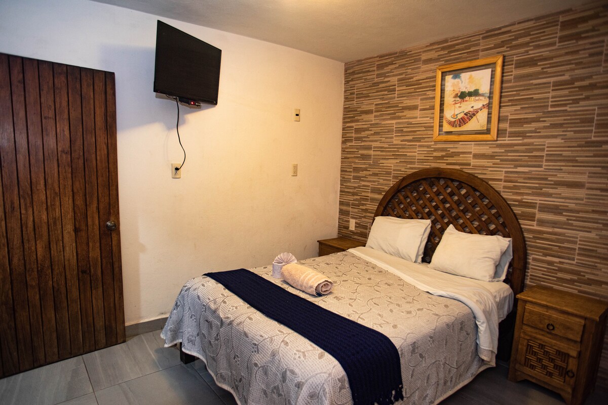 A cozy room is furnished with a spacious bed featuring a decorative headboard. A television is mounted on the wall opposite the bed. Soft lighting complements the textured wallpaper, while a piece of artwork adds a touch of character to the space.