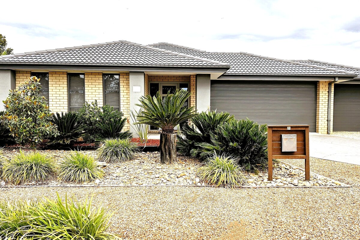 The exterior of a newly built house is shown, featuring brick walls and a tiled roof. Lush landscaping with pathway stones, flowering plants, and shrubs enhances the entrance. A modern mailbox stands beside the driveway, which leads to a spacious triple garage.