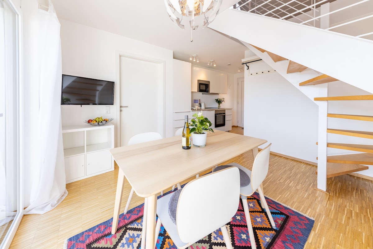 A dining area features a wooden table surrounded by white chairs, with a potted plant as the centerpiece. Natural light pours in from a nearby window. In the background, a modern open kitchen can be seen, along with a staircase leading to the upper level.