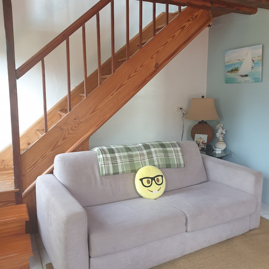 A light-colored sofa with a playful pillow is positioned beside a small lamp and a piece of wall art. The staircase with wooden railings leads to the upper level, and soft natural light is present in the space.