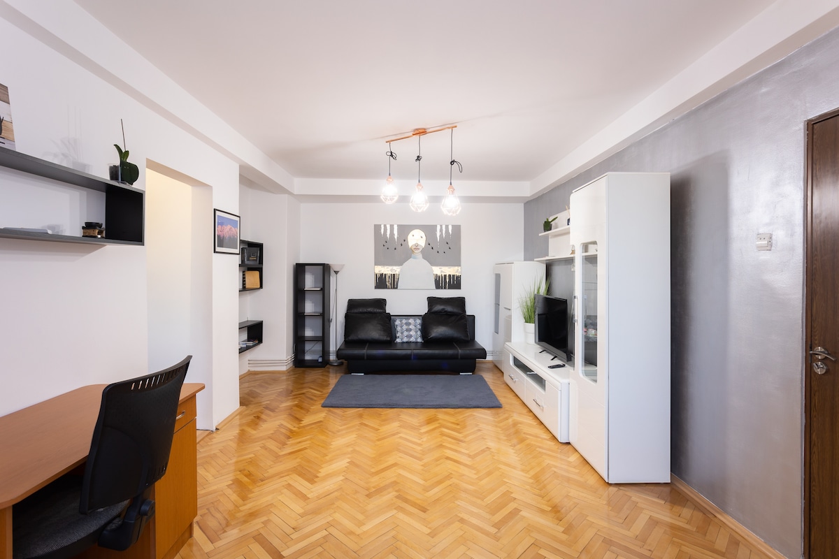 The living room features a modern black sofa facing a wall-mounted TV unit. A wooden desk with an office chair is positioned to one side. Decorative shelving and a stylish light fixture add a contemporary touch. Light-colored walls and wooden flooring create a warm environment.