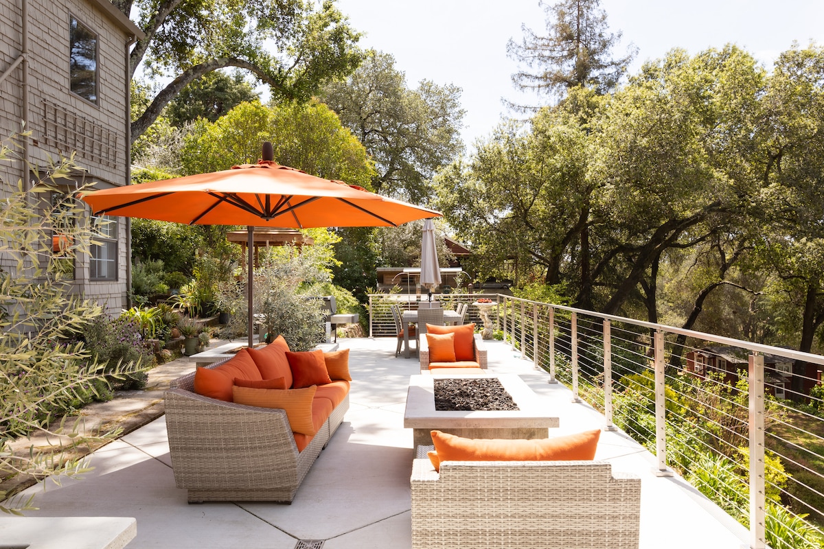 An outdoor patio area is shown, featuring comfortable seating with orange cushions and an orange umbrella providing shade. A table with a fire pit is positioned nearby, surrounded by greenery and trees, creating a relaxing environment for outdoor leisure.