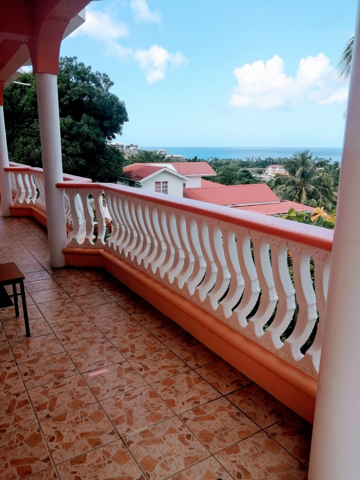 A balcony is featured with a tiled floor, showcasing a railing with arches and a view of the nearby neighborhood and distant ocean. Lush greenery is visible in the foreground, and the sky is bright with scattered clouds.