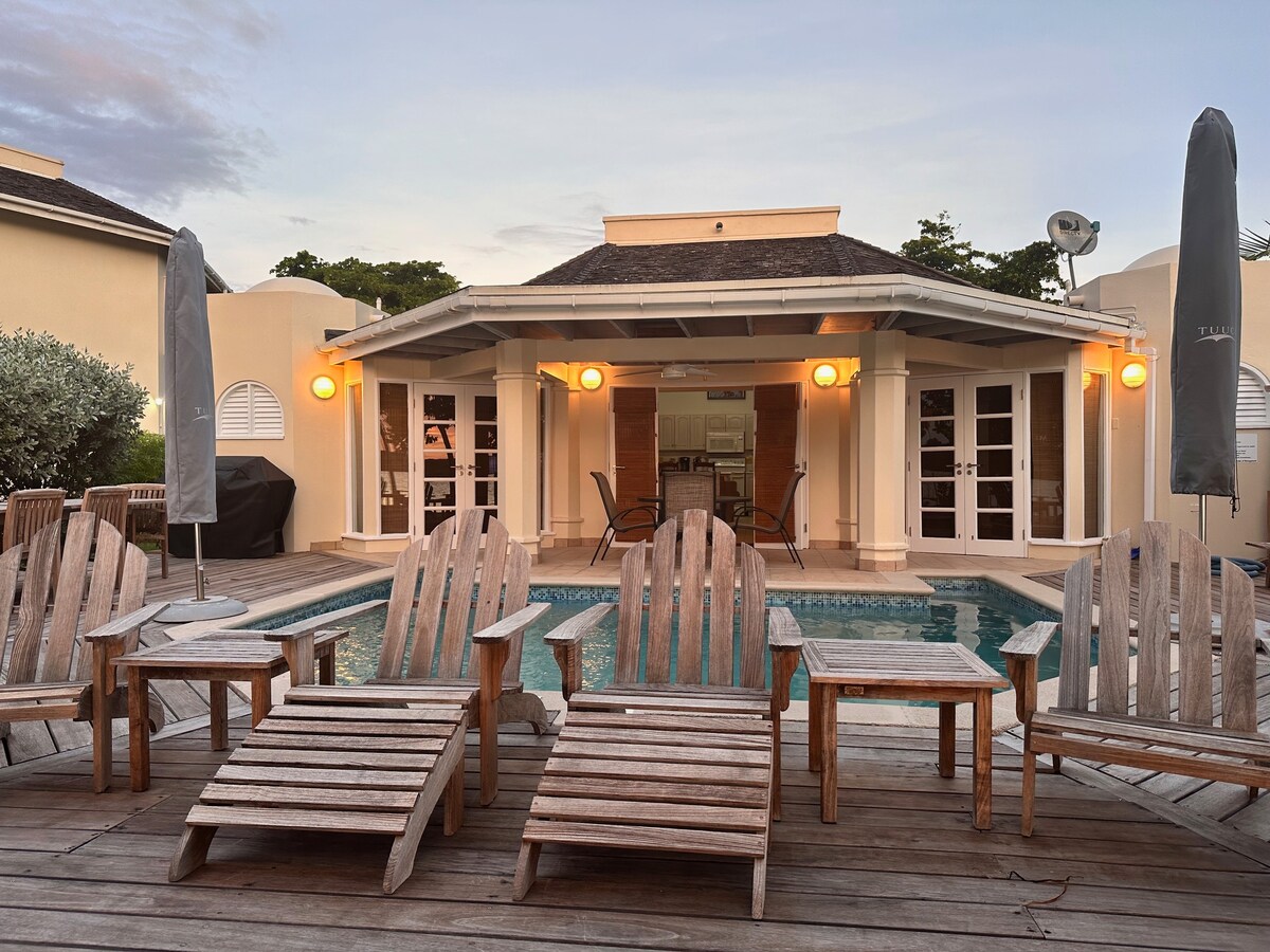 A collection of wooden lounge chairs is arranged on a deck near a pool. The bungalow features large glass doors that open to the pool area, with outdoor seating visible in the background. Soft lighting creates a welcoming ambiance as the sun sets.