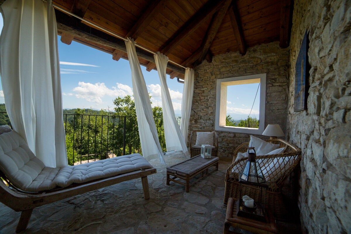 A cozy terrace features a wooden lounge chair and a wicker seating arrangement, complemented by a large lantern. Sheer white drapes flow gently in the breeze, framing a stunning view of the surrounding greenery and distant mountains through a large window.