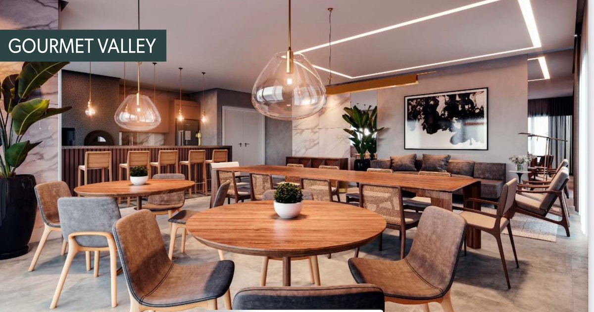 A modern dining area features multiple wooden tables surrounded by varied seating options. Soft lighting fixtures hang from the ceiling, highlighting the warm tones of the furniture. A central green plant adds a touch of nature to the stylish space, which is designed for communal gatherings.