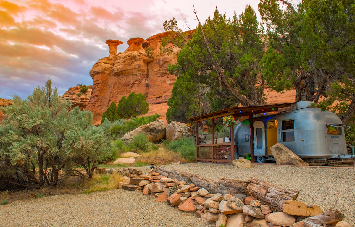 A refurbished 1958 Airstream is set against a backdrop of striking red rock formations. The outdoor area features a gravel space bordered by natural stone, with a covered patio that offers shade and a cozy ambiance, complemented by greenery from nearby trees.