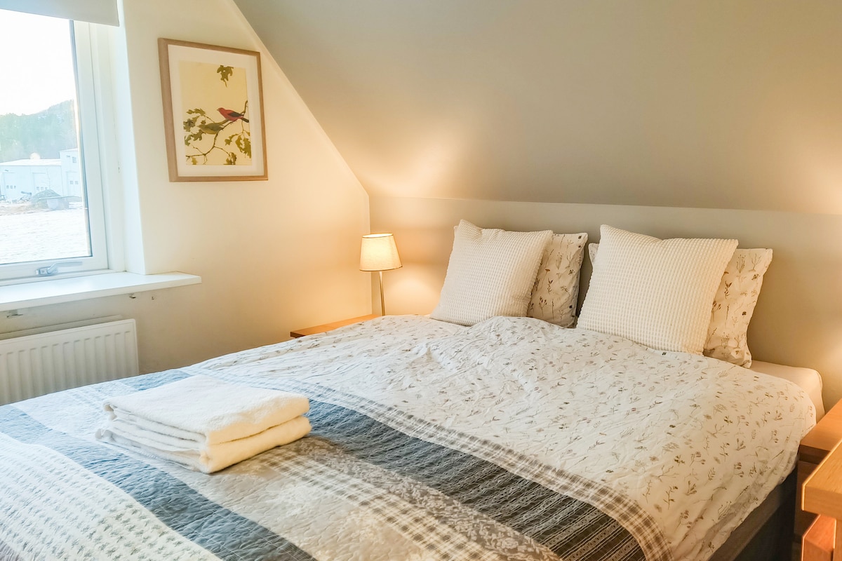 A spacious bed with a patterned duvet and soft pillows is illuminated by gentle bedside lamps. Natural light filters in through a nearby window, enhancing the serene ambiance. A piece of artwork featuring a bird is displayed above the bed, adding a subtle decorative touch.