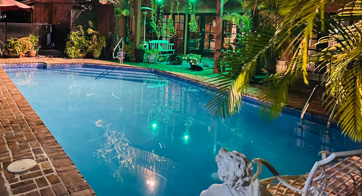 A serene swimming pool reflects soft green lights at night, surrounded by lush tropical plants and brick pathways. Comfortable seating is positioned nearby, enhancing the inviting atmosphere of the outdoor area. The space is designed for relaxation and enjoyment.