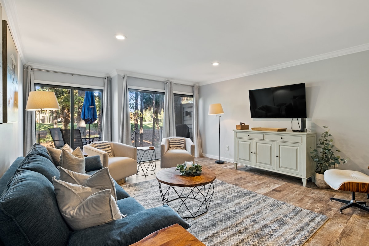 The living room features a cozy sectional sofa paired with two accent chairs, arranged around a circular coffee table. Natural light flows in through large sliding doors, leading to a patio with views of the surrounding greenery. A smart TV is mounted on the wall.