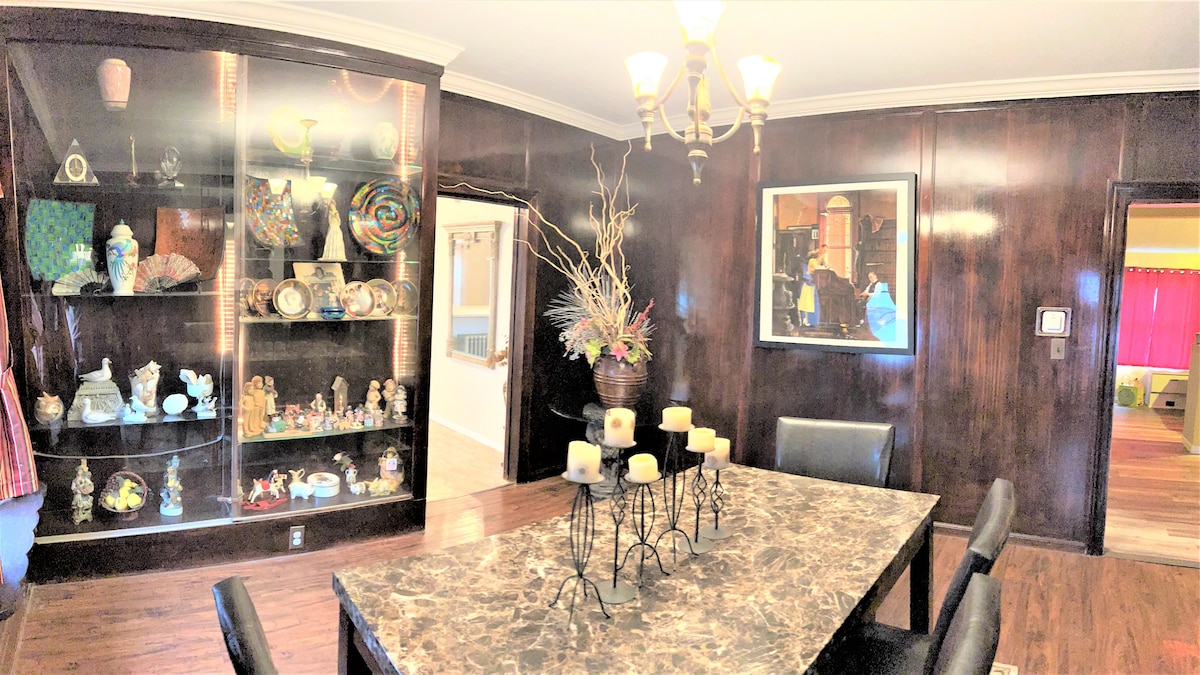 A dining area with a dark wood-paneled wall features a large marble table surrounded by black chairs. A display cabinet showcases colorful decorative items and quirky artwork, enhancing the space's character. Soft lighting from a chandelier adds illumination to the room.