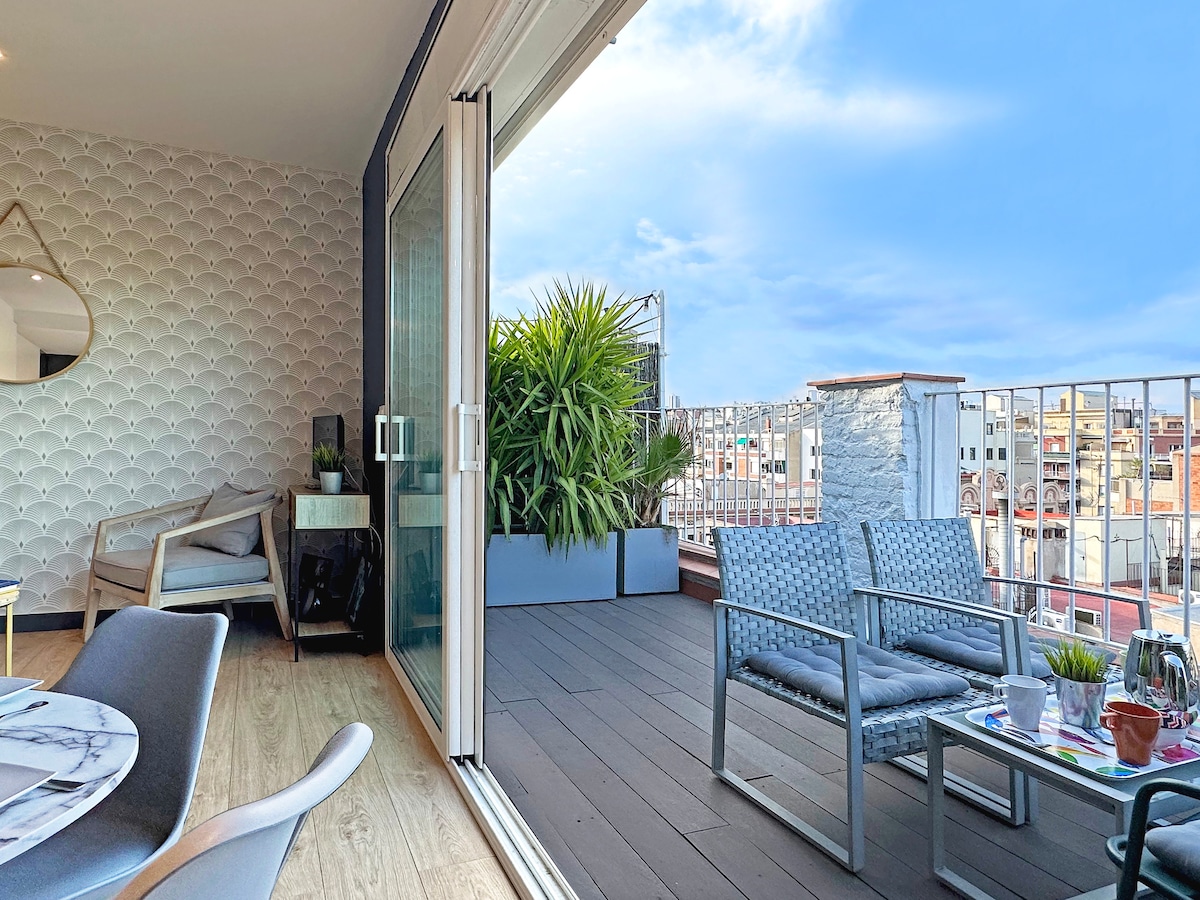 A spacious terrace is visible, featuring contemporary outdoor furniture, including two armchairs and a small table. Lush greenery adds a natural touch, while large sliding glass doors provide access from the interior, allowing for an open-air atmosphere.