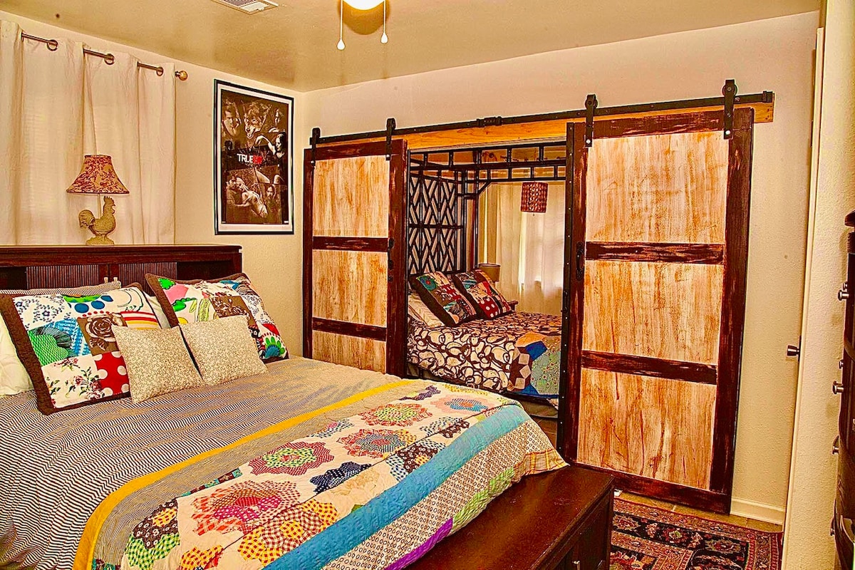 A bedroom features a spacious bed adorned with a vibrant quilt, layered with decorative pillows in various patterns. A unique sliding door system separates another sleeping area with a playful bunk bed design. Soft curtains frame a window, allowing natural light to enter the room.