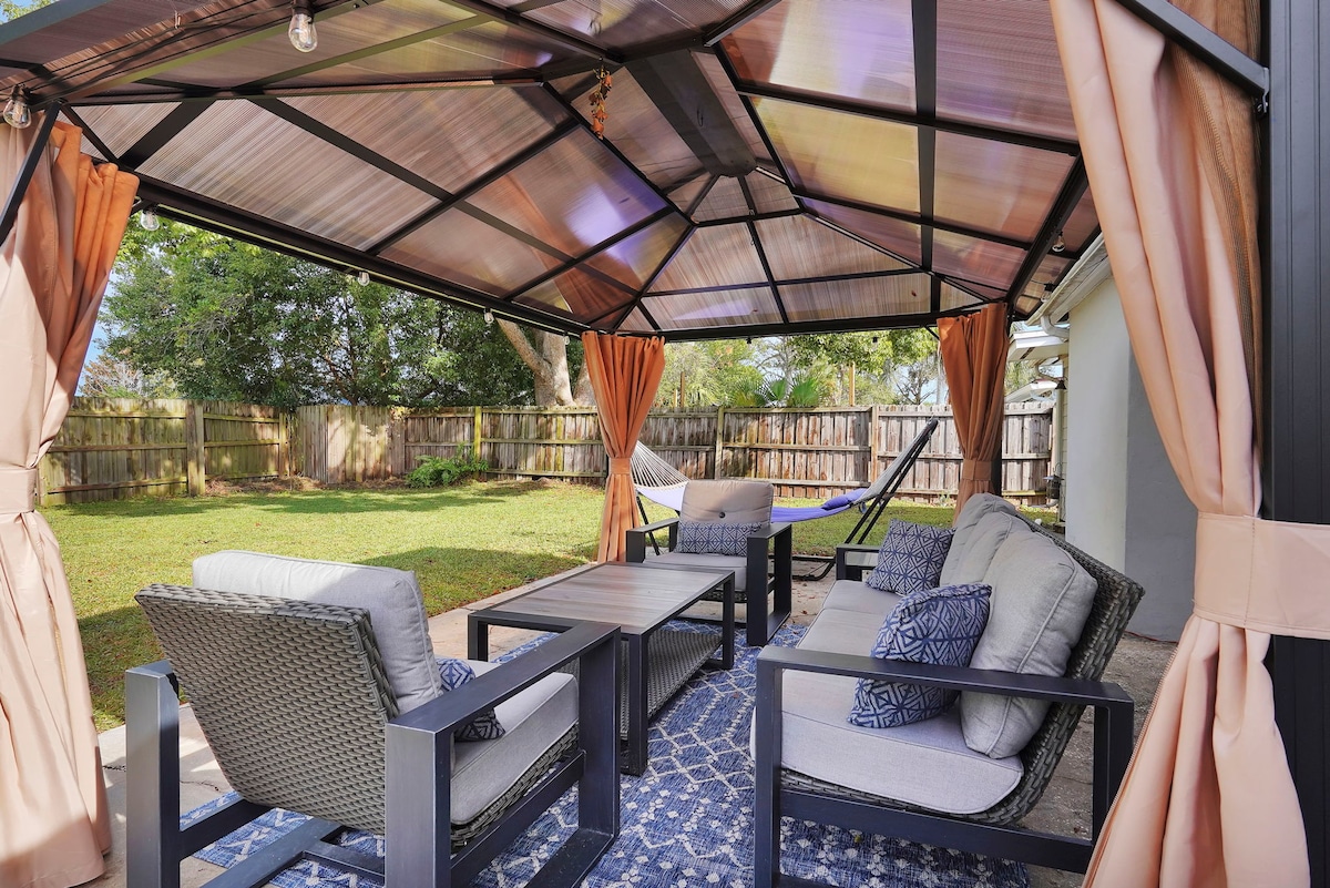 An outdoor seating area is sheltered by a canopy, featuring comfortable chairs with soft cushions arranged around a simple table. Orange drapes frame the space, and a hammock is visible in the background against a lush green lawn.