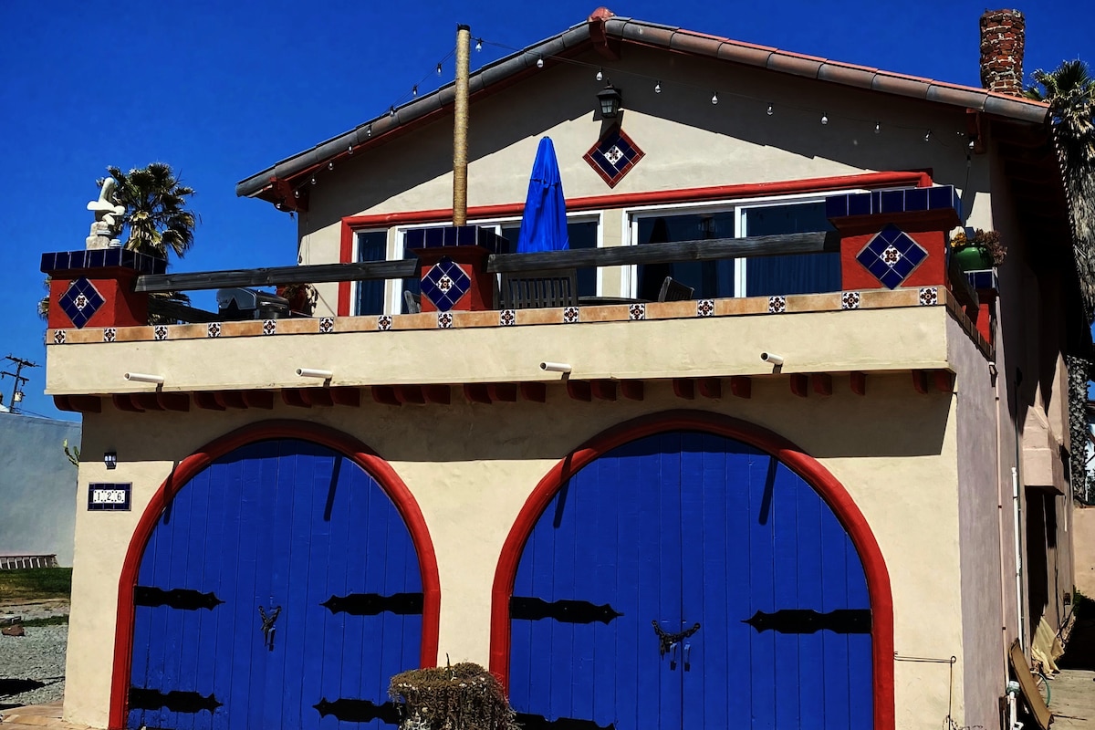 The exterior of the beach home is adorned with colorful tiles and features a distinct two-car garage with vibrant blue doors. A second-floor deck is visible, equipped with a blue umbrella for shade, providing an inviting outdoor space.