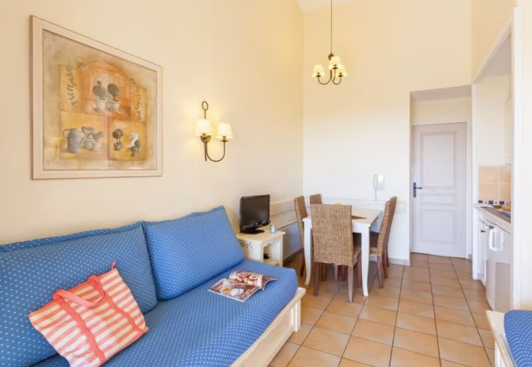 A comfortable living area features a blue sofa adorned with cushions, a small television, and a dining table surrounded by wicker chairs. The walls are painted in a soft hue, complemented by a piece of artwork and lighting fixtures that enhance the ambiance.