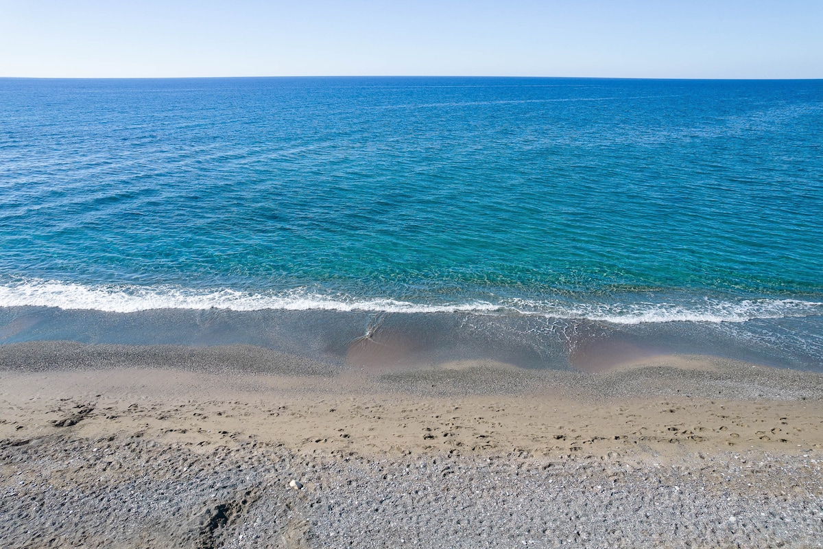 A serene beach scene features gentle waves lapping at a smooth sandy shore. The clear blue waters reflect the sky, while a subtle gradient can be seen where the ocean meets the beach. The sandy area is dotted with small pebbles, adding to the natural ambiance.