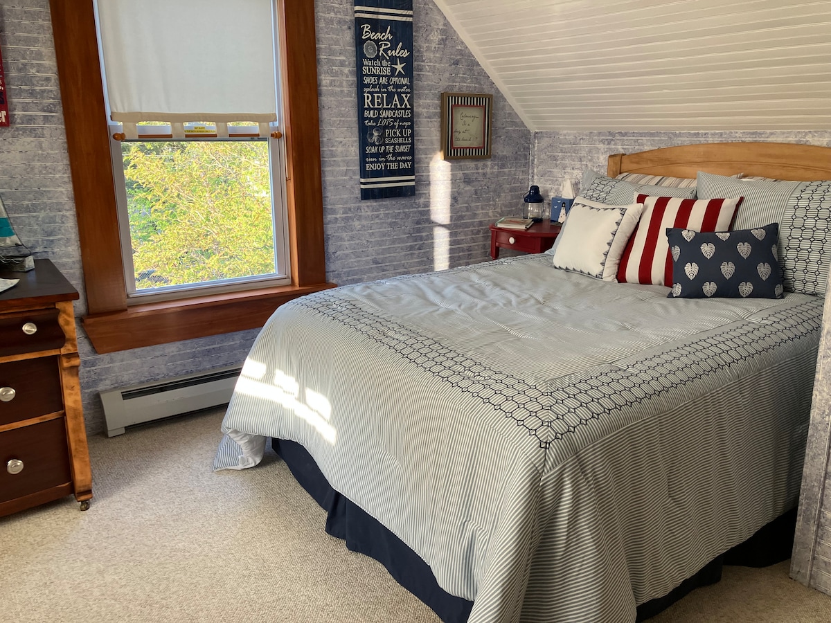 A light-filled bedroom features a cozy bed dressed in a patterned blue and white quilt. A decorative arrangement of red and navy pillows enhances the inviting look. Sunlight streams through a window, illuminating the soft beige carpet and accentuating the room's charming decor.