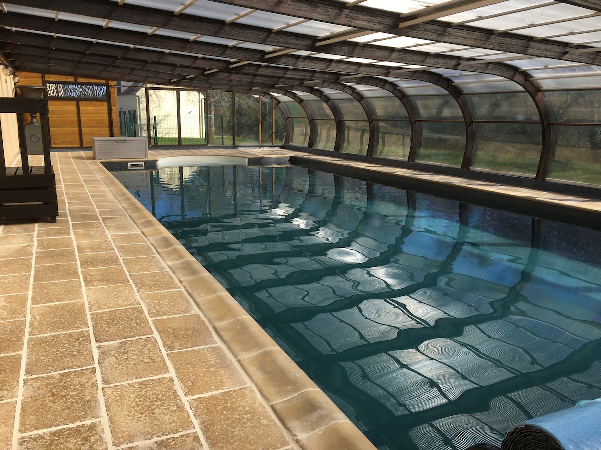 An indoor pool area features a clear, covered ceiling that allows natural light to illuminate the water. The spacious pool is framed by a textured tiled floor, enhancing the tranquil atmosphere. The glass sides provide views of the surrounding greenery.