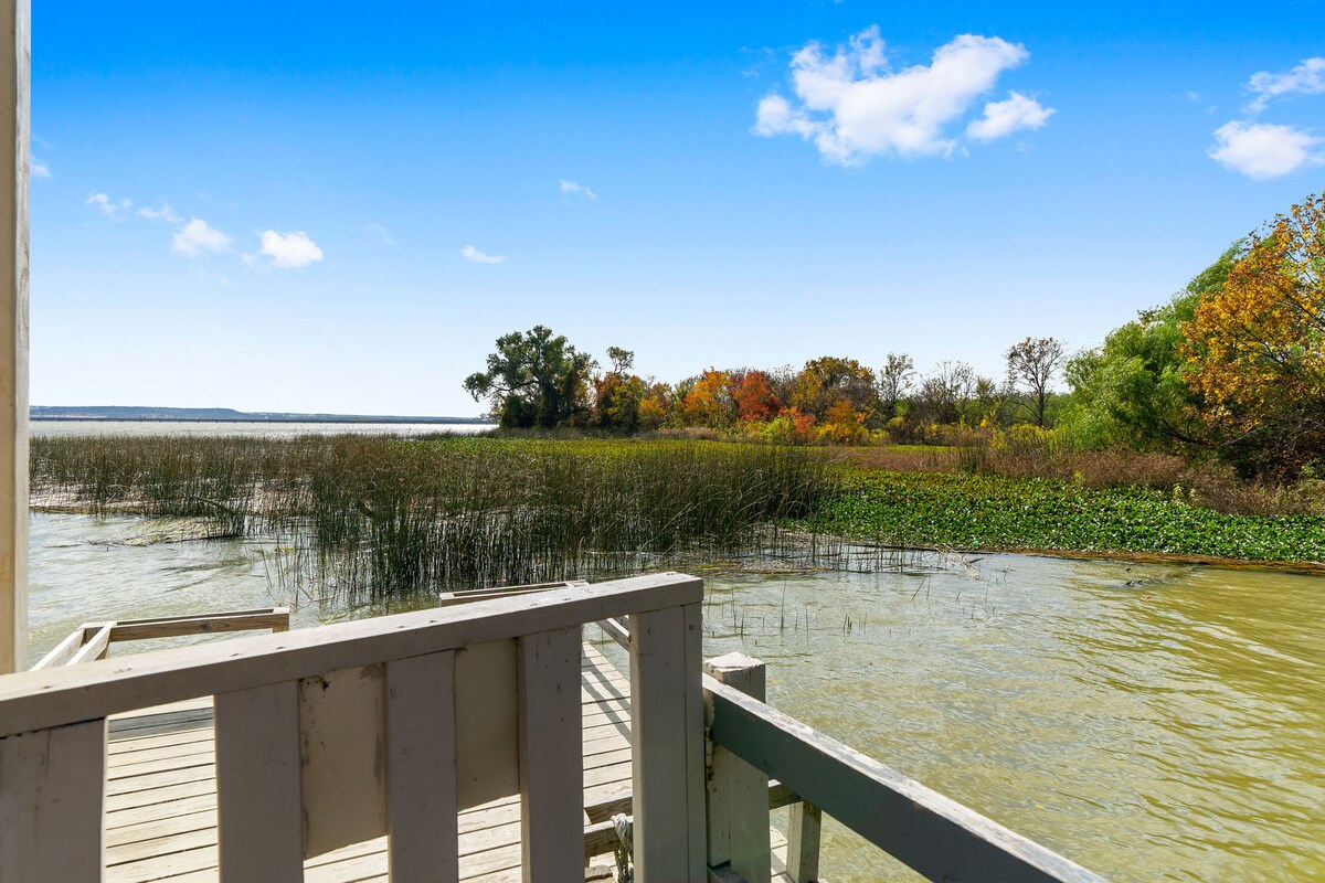 A serene view of the lake is captured from the dock, showcasing calm waters reflecting the sky. Nearby, lush greenery and reeds frame the scene, with trees showing autumn colors in the background. The peacefulness of nature is emphasized by the open setting.