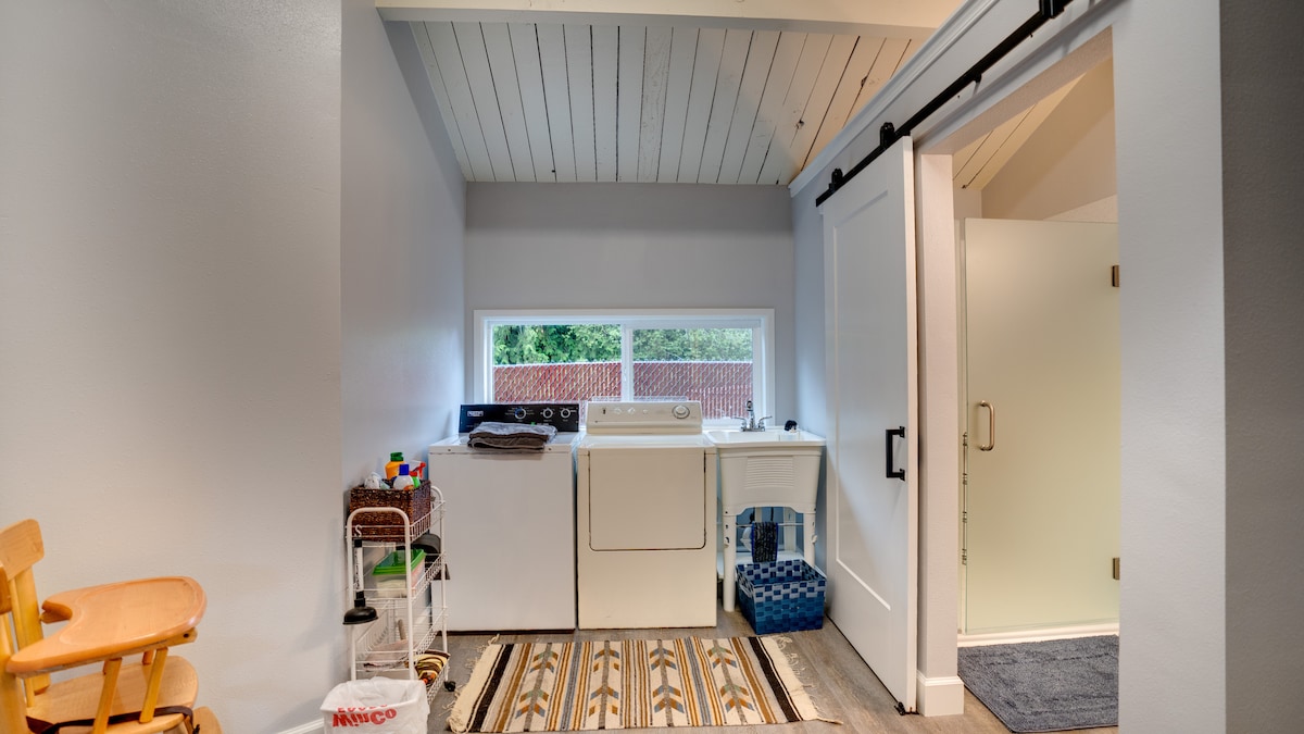 A laundry area is observed with a washing machine and dryer side by side. A large window allows natural light to brighten the space, revealing a sliding barn door. A patterned rug runs along the floor, complemented by a small chair and organized shelving for supplies.