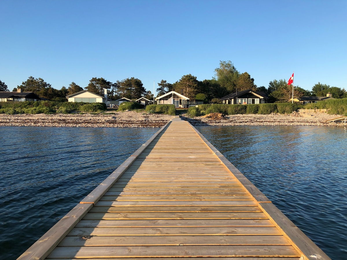 A wooden dock extends over calm waters towards a waterfront property. The scene includes several low-profile buildings surrounded by trees. A flag is visible, indicating the property's location. The sky is clear, suggesting a serene and inviting atmosphere.