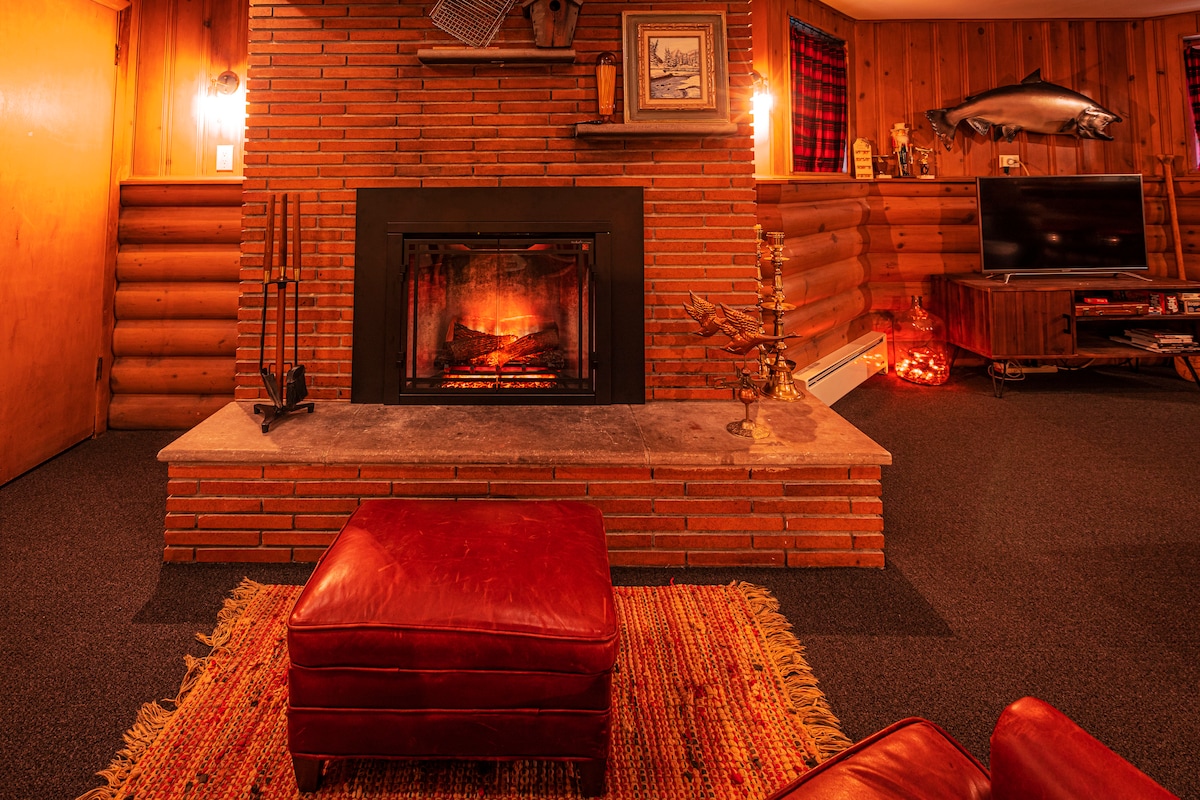 A rustic fireplace is set in a brick wall, surrounded by a spacious area featuring a vibrant area rug. A cozy ottoman rests nearby, with warm light radiating from wall sconces. A television is positioned on a wooden entertainment unit opposite the fireplace.