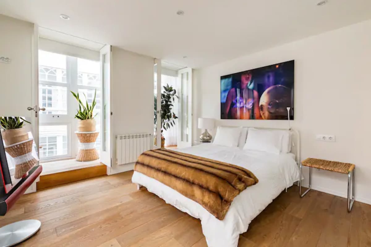 A bright bedroom features a spacious bed adorned with a soft blanket. Natural light floods the room through large windows, accompanied by potted plants. A modern artwork hangs above the bed, and a small woven stool is positioned beside it, enhancing the room's functionality.
