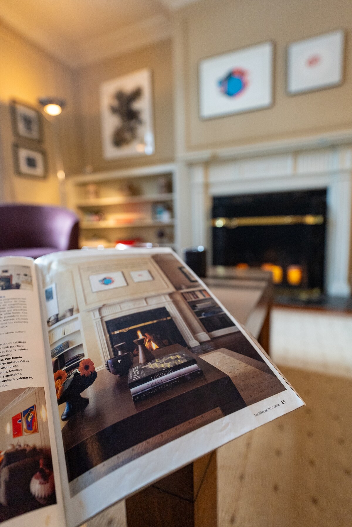 A living room is visible with warm tones enhancing the ambiance. In the foreground, an open magazine lies on a wooden coffee table, featuring a fireplace. The wall displays framed art pieces and a tasteful bookshelf, which adds character to the space.
