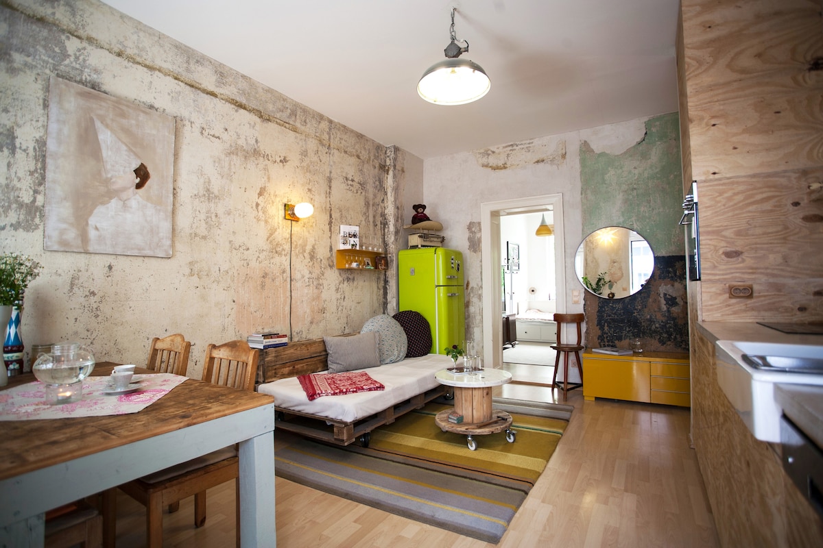 A stylish living area features a mix of vintage furniture with a rustic table, a sofa bed enhanced by colorful textiles, and an eye-catching bright green refrigerator. The walls showcase a blend of textured plaster, while a round mirror and art add visual interest to the space.