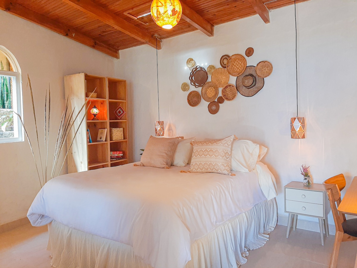 A bright bedroom features a spacious bed with crisp white linens and decorative pillows. Woven wall art adds a touch of texture, while bedside lamps provide a soft glow. A wooden bookshelf and a small desk create functional spaces, with natural light illuminating the room.
