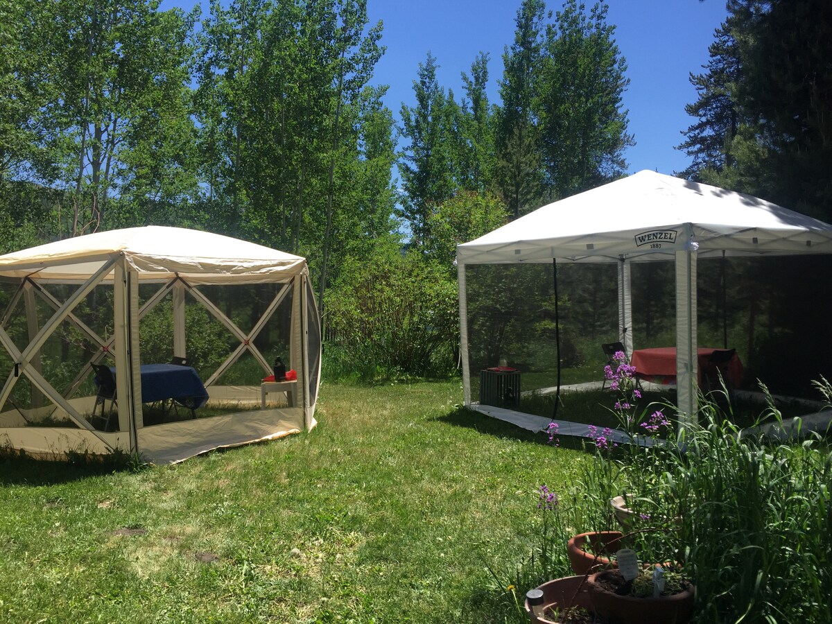 Two outdoor tents are positioned on a grassy area, surrounded by dense greenery. One tent is screened, providing an open space for dining, while the other is a white pop-up structure with visible tables set for guests. Colorful wildflowers bloom nearby.