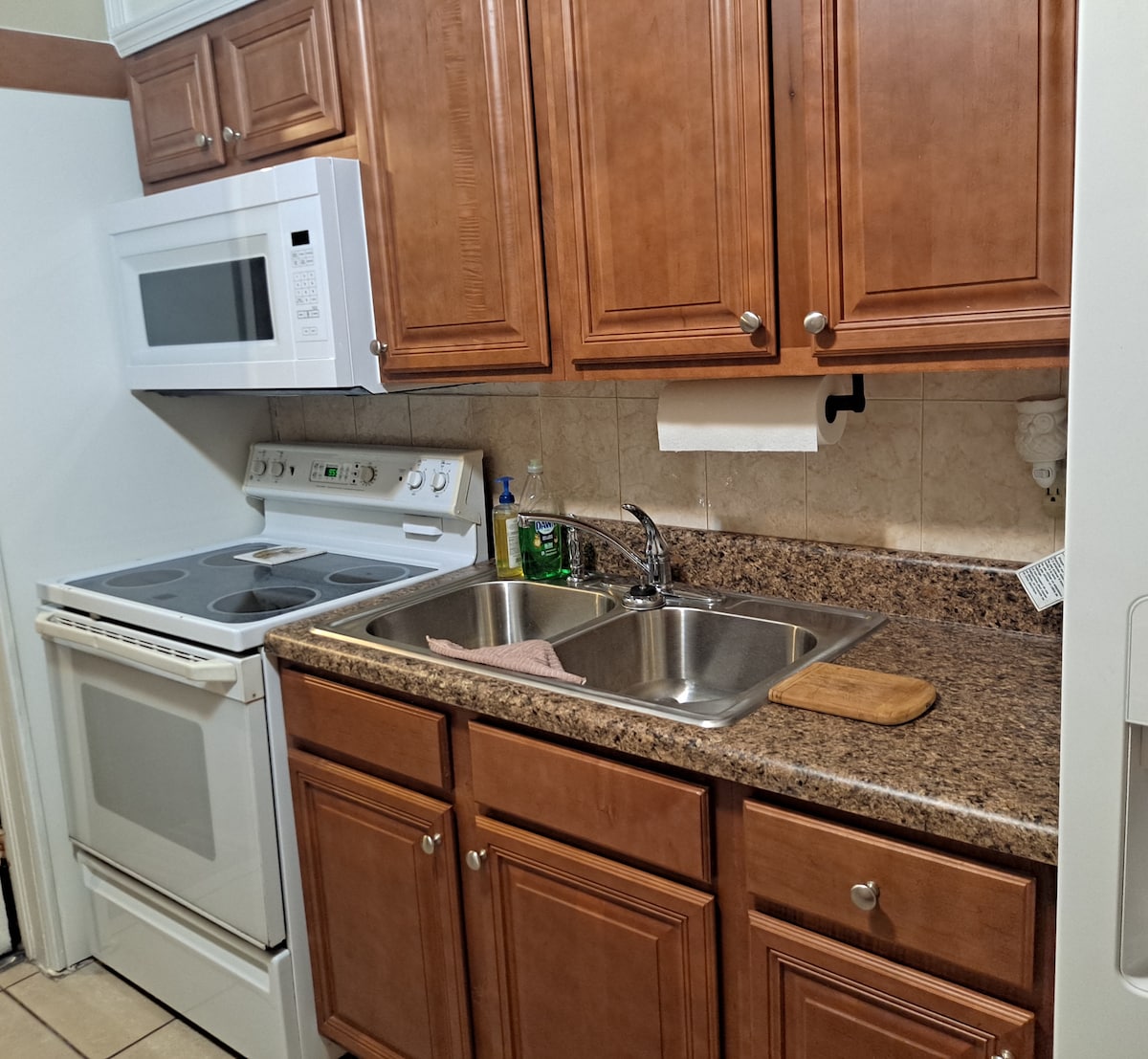The kitchen features wooden cabinetry with a warm finish, equipped with a stove, microwave, and double sink. A granite countertop offers a functional space for meal preparation, with basic supplies visible for convenience. Tile flooring enhances the kitchen's practical layout.