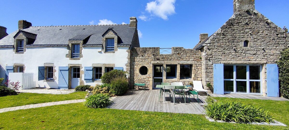 The exterior of a stone house is showcased in a lush green setting. A wooden deck features a dining table surrounded by chairs, while large windows invite natural light. The charming mix of traditional and modern architecture is complemented by a blue sky and scattered clouds.