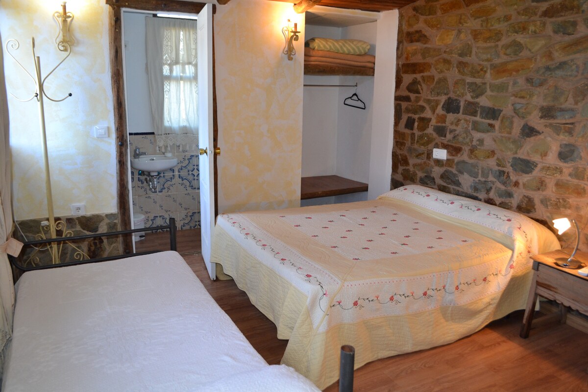 A cozy room features a spacious bed along with an additional single bed, both adorned with light-colored bedding. Exposed stone walls add rustic charm, while a small closet area provides storage. A private bathroom is visible through an open door, enhancing convenience.