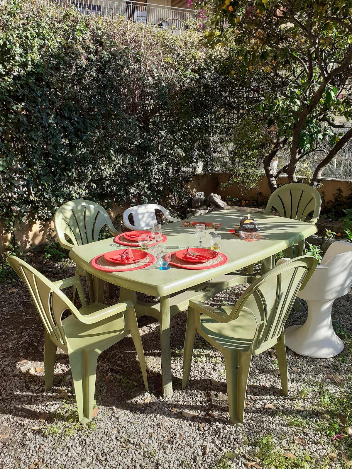 An outdoor dining area features a green table surrounded by five matching chairs. Plates and glassware are set on the table, ready for a meal. Natural sunlight filters through surrounding foliage, creating a pleasant outdoor ambiance.