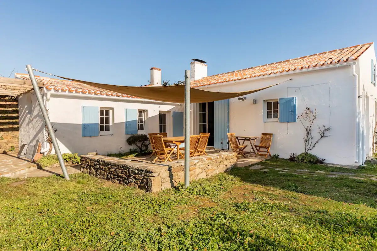 A traditional white cottage features blue shutters, sitting under a clear sky. A shaded terrace is equipped with wooden chairs and a table, positioned on a grassy area. The stone pathway leads to the entrance, enhancing the inviting outdoor space.