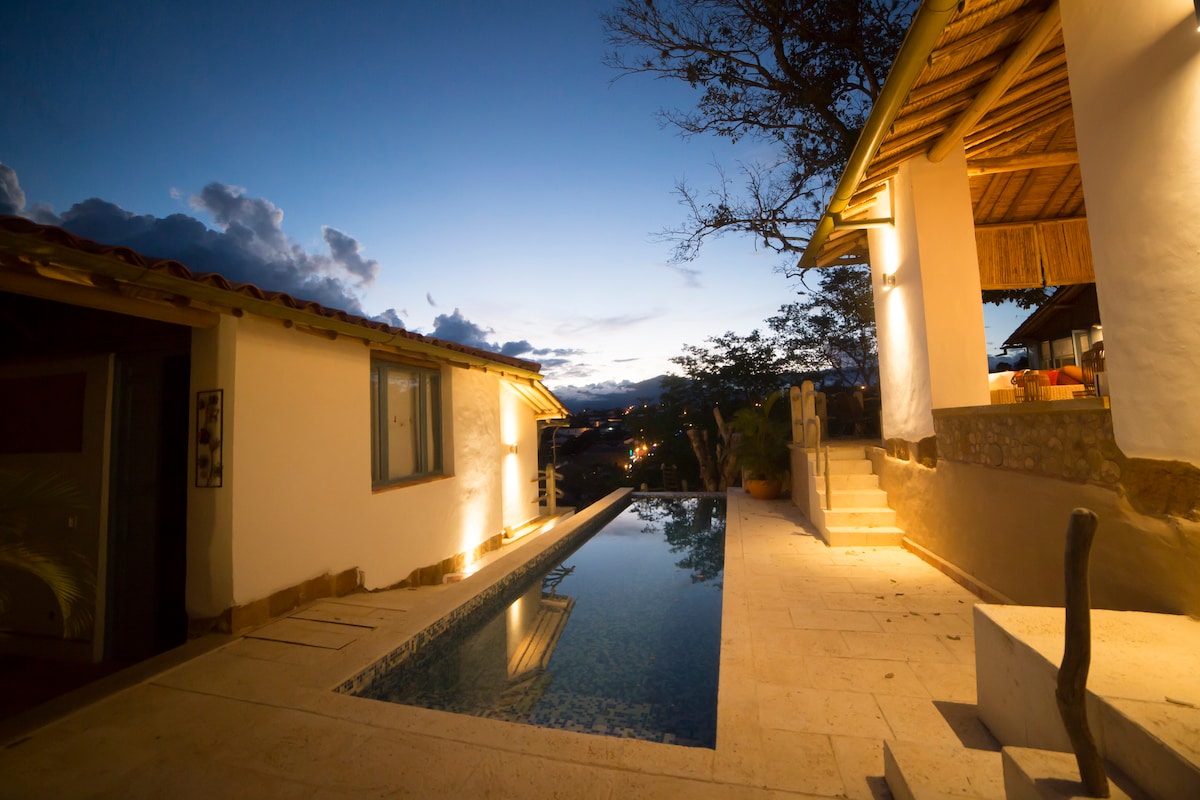 A serene evening scene features a swimming pool reflecting the twilight sky. The nearby structure, with a thatched roof, is softly illuminated. A stone pathway leads from the pool to an inviting entrance, enhancing the outdoor living space.