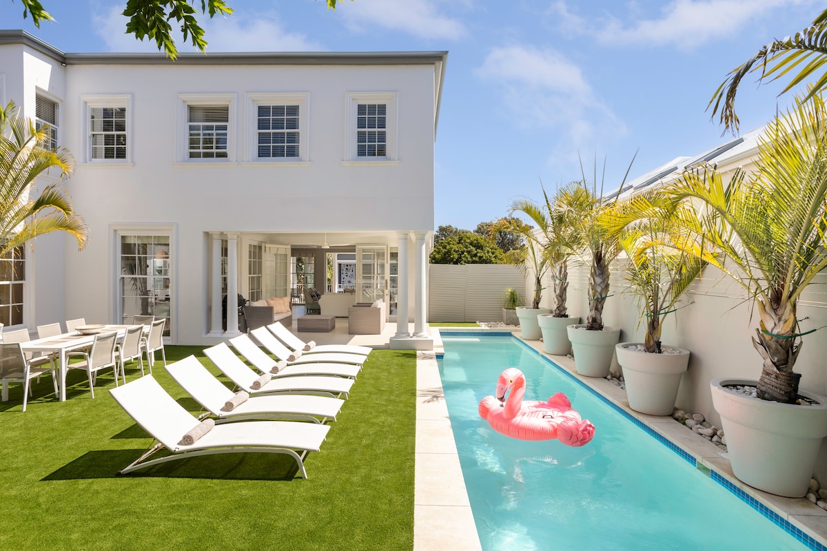 A sunny courtyard features a lap pool surrounded by palm trees and planters. Multiple lounge chairs are arranged neatly on green grass, alongside a dining table and chairs. A pink inflatable float adds a playful touch to the pool, creating a relaxing outdoor space.