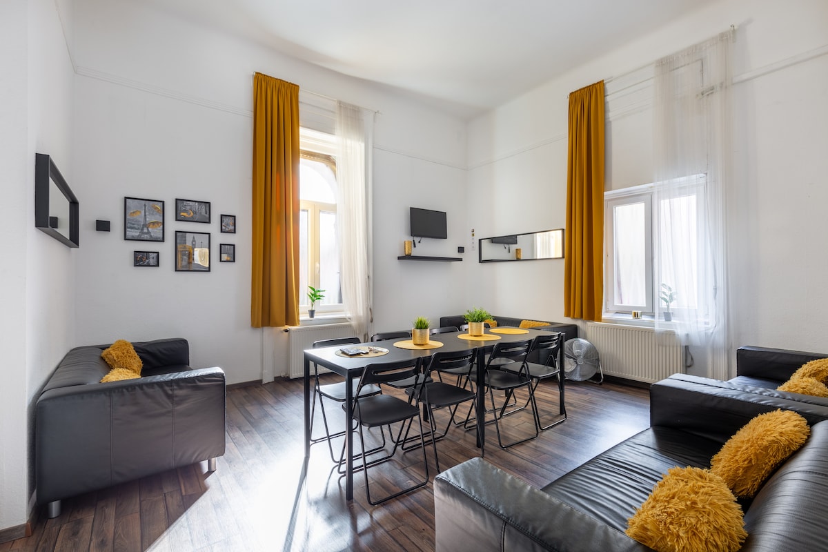 A spacious living area is furnished with two black sofas and a dining table set for ten. Tall windows dressed with light curtains allow natural light to fill the room. Decor includes framed artwork on the walls and a mirrored surface, creating a bright and welcoming ambiance.