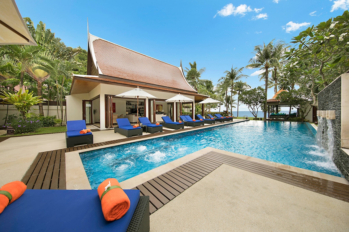 An expansive swimming pool is surrounded by comfortable lounge chairs and umbrellas, set against a backdrop of lush greenery and palm trees. The villa features a traditional Thai architectural style, with open living spaces visible in the distance and the serene Gulf of Thailand beyond.