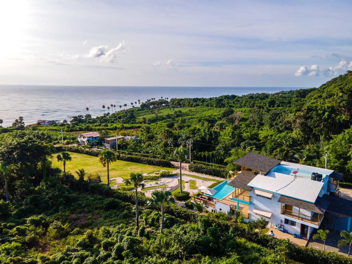 An aerial view captures a private villa set among lush greenery, with a sparkling pool reflecting the sky. The property transitions into a vast landscape of trees and palms leading to the coastline, where the gentle waves of the ocean can be seen in the background.