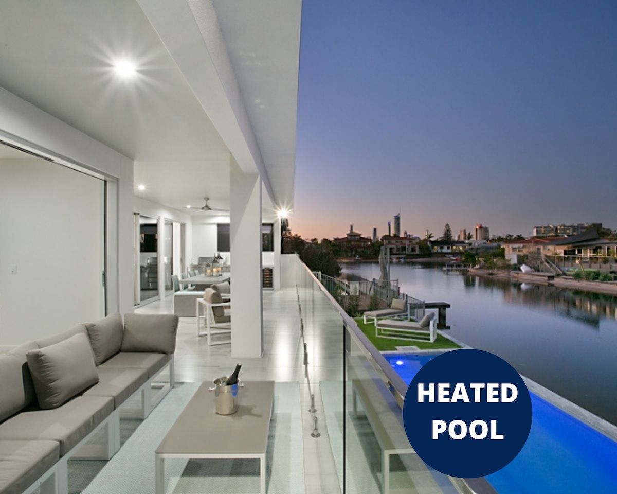 An expansive outdoor terrace offers comfortable seating with a view of the water at dusk. The heated pool glows in vibrant blue, while the distant skyline of Broadbeach provides an appealing backdrop. Glass railings enhance the open feel of the space.