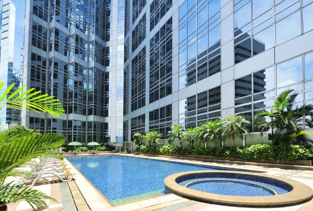 A modern outdoor swimming pool is surrounded by tall glass buildings, reflecting natural light. Lounge chairs line the poolside, and tropical plants create a serene atmosphere. A circular hot tub is adjacent to the pool, offering a place to relax.