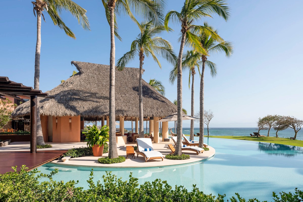 A thatched roof palapa is surrounded by swaying palm trees and turquoise waters, creating a serene outdoor space. Lounge chairs are positioned around a gently curving pool, offering views of the coastline and lush landscaping, enhancing the connection to the natural environment.