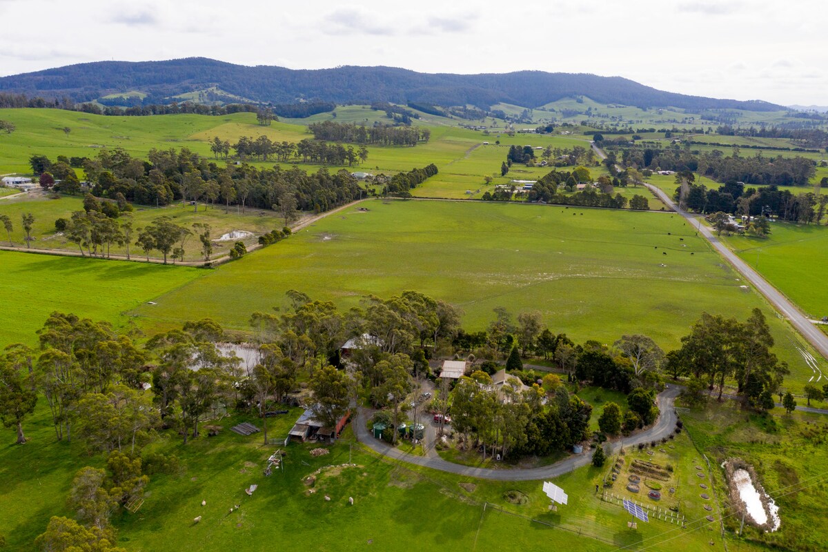 Aerial view of a vast green landscape featuring rolling hills and a variety of trees. The property includes a garden area and a winding driveway leading to the unit, surrounded by open fields and farmlands.