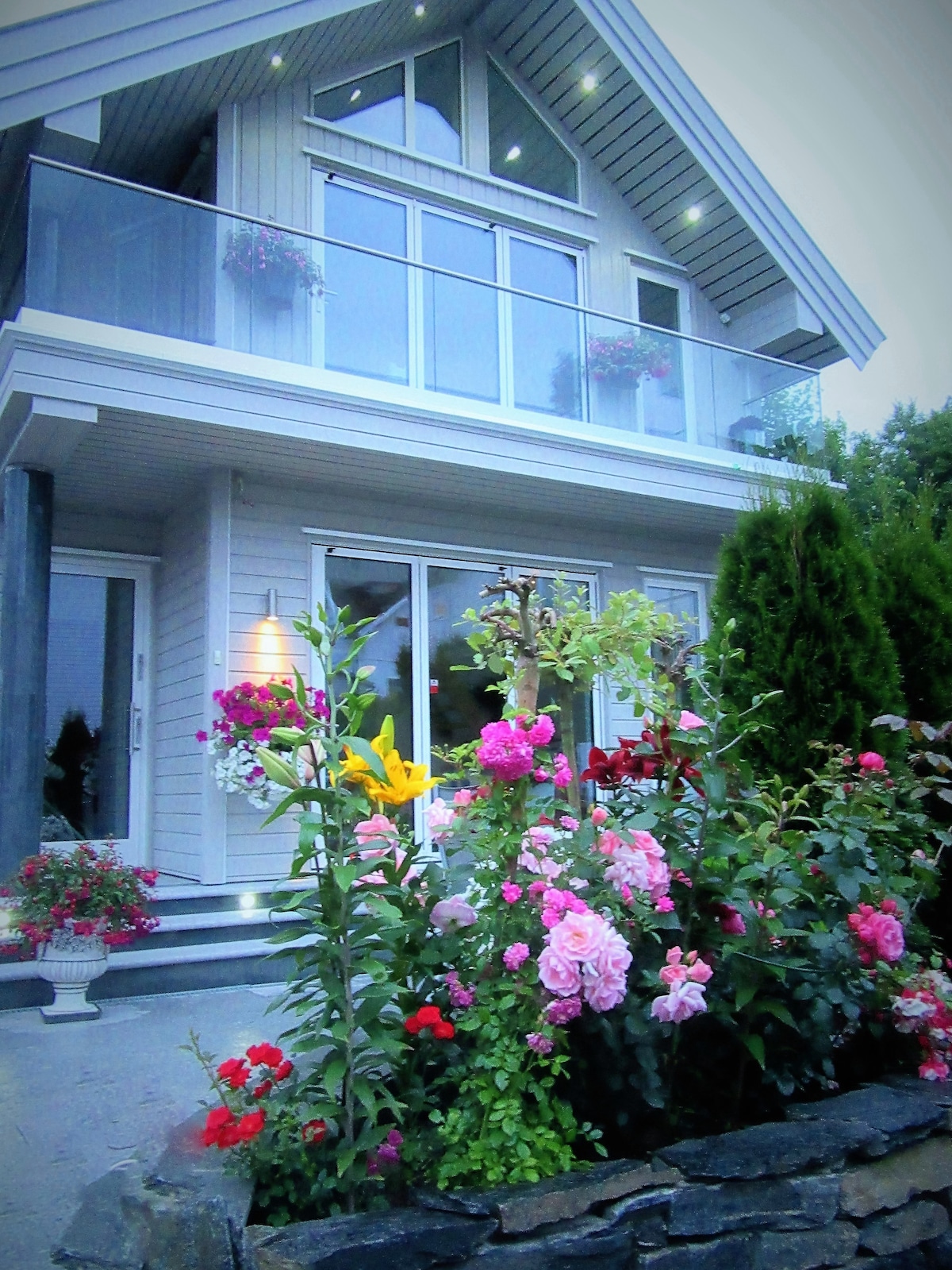 The exterior of a modern house showcases a sloped roof and large glass windows, allowing ample natural light. Colorful flowers, including roses and lilies, adorn the garden, while potted plants are located near the entrance, enhancing the outdoor setting.