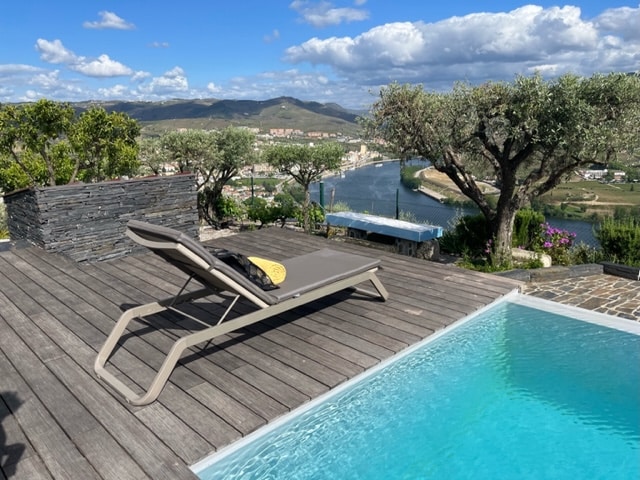 A loung chair is positioned beside a clear pool, with views of the Douro River and surrounding vineyards. Olive trees provide shade in the landscaped area, while a stone wall adds structure to the outdoor setting against a backdrop of rolling hills and blue skies.