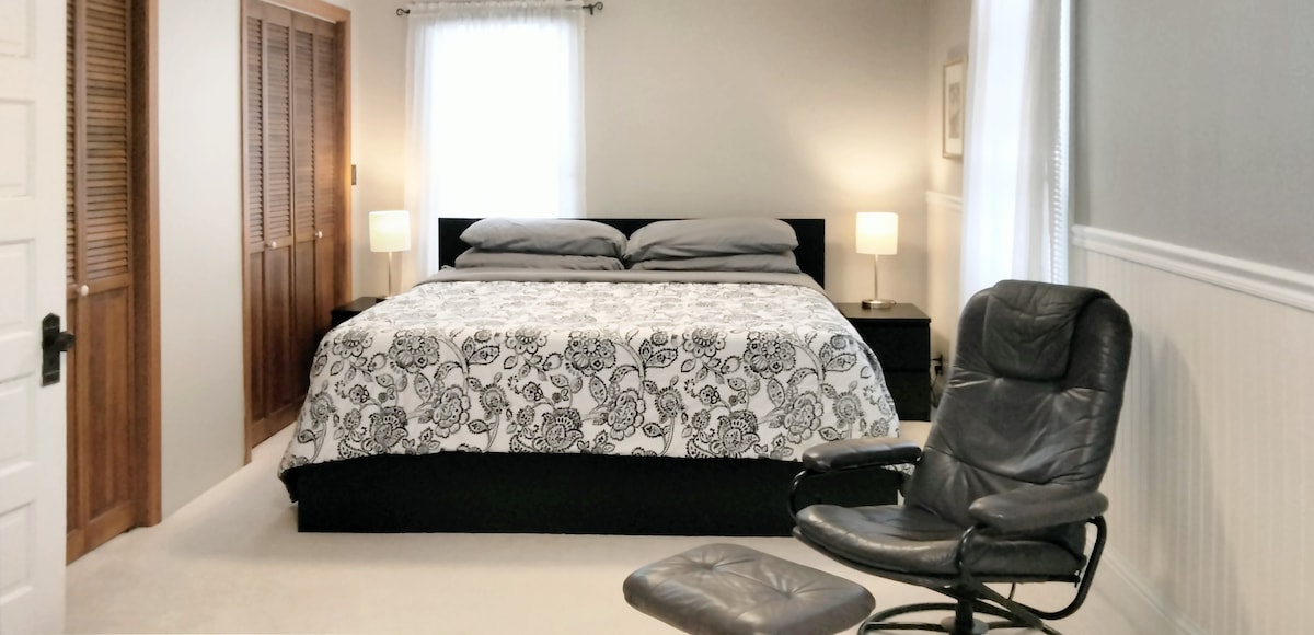 The bedroom features a spacious bed with a patterned comforter and two nightstands, each topped with a lamp. A comfortable leather chair is positioned nearby. Natural light enters through two windows with sheer curtains, complemented by light-colored walls and a cozy carpeted floor.