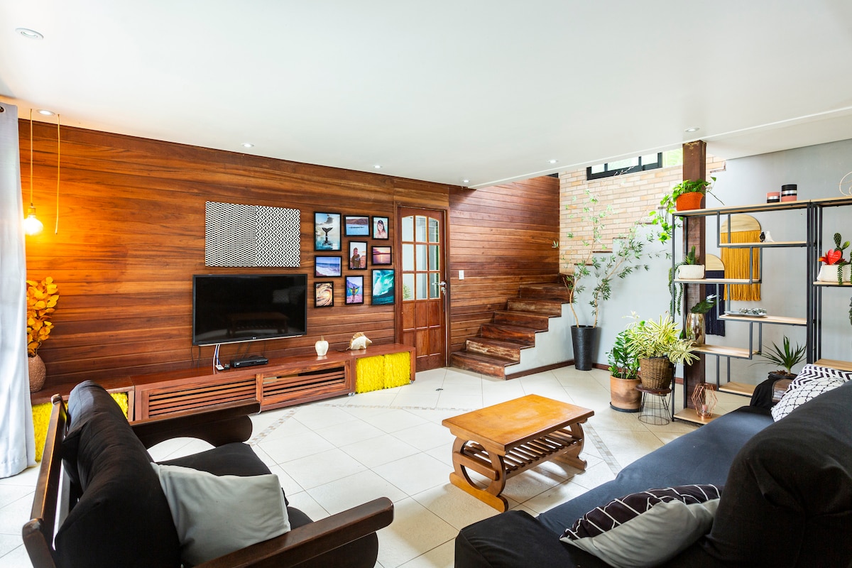 A spacious living area features comfortable black sofas arranged around a wooden coffee table. A flat-screen TV is mounted on a wooden entertainment unit. Natural light illuminates the space, with decorative plants and framed artwork enhancing the inviting environment.